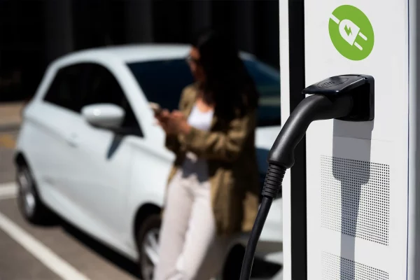 Nahaufnahme einer Ladesäule mit eingestecktem Ladekabel, im Hintergrund unscharf eine Person neben einem weißen Elektroauto.