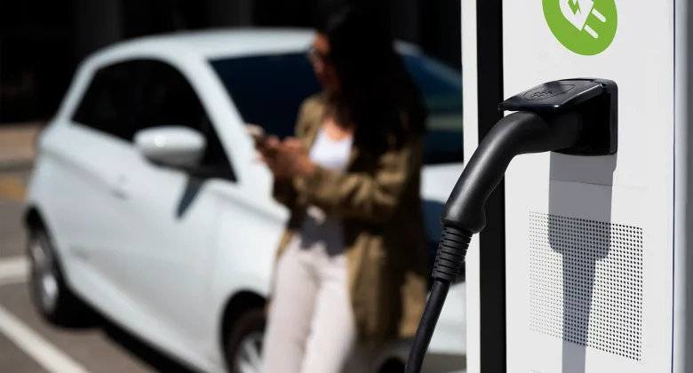 Nahaufnahme einer Ladesäule mit eingestecktem Ladekabel, im Hintergrund unscharf eine Person neben einem weißen Elektroauto.